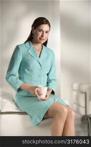 Caucasian mid adult professional business woman sitting in modern office holding coffee mug looking at viewer and smiling.