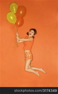 Caucasian mid-adult woman being lifted into the air by balloons, on orange background.