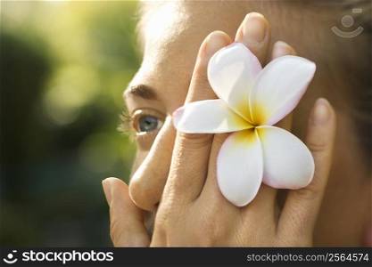 Caucasian mid-adult woman holding plumeria flower over eye.