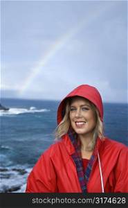 Caucasian mid-adult woman in red raincoat in front of ocean with rainbow in background looking at viewer and smiling in Maui, Hawaii.