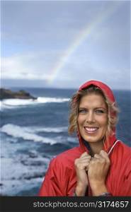 Caucasian mid-adult woman in red raincoat standing by ocean with rainbow in background in Maui, Hawaii.