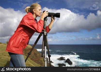 Caucasian mid-adult woman looking through camera on tripod on cliff overlooking ocean in Maui, Hawaii.