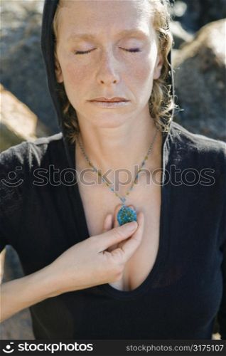 Caucasian mid-adult woman wearing black hoodie holding pendant necklace with eyes closed.