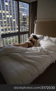 Caucasian mid-adult woman wearing lingerie lying in bed looking out window at cityscape.