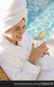 Caucasian mid-adult woman wearing robe and towel on head drinking from glass next to pool.