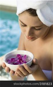 Caucasian mid-adult woman wearing towel around head and body holding bowl of purple orchids next to pool.