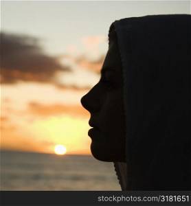 Caucasian mid-adult woman with scarf over head profiled against sunset over ocean.