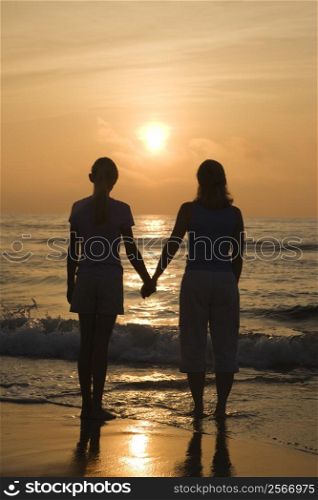 Caucasian mid- female and teenage daughter standing on beach at sunset holding hands.