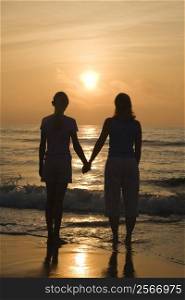 Caucasian mid- female and teenage daughter standing on beach at sunset holding hands.