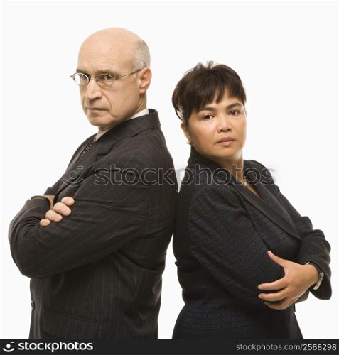 Caucasian middle-aged businessman and Filipino businesswoman standing back to back with arms crossed looking serious.