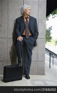 Caucasian middle aged businessman leaning on wall outdoors with foot on briefcase.