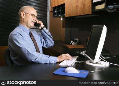Caucasian middle-aged businessman sitting at desk in office talking on telephone and typing on computer.