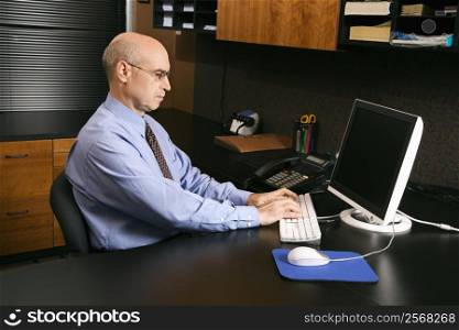 Caucasian middle-aged businessman sitting at desk in office typing on computer.