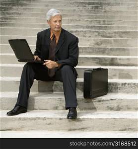 Caucasian middle aged businessman sitting on steps outdoors with laptop and briefcase.