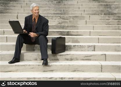 Caucasian middle aged businessman sitting on steps outdoors with laptop and briefcase.