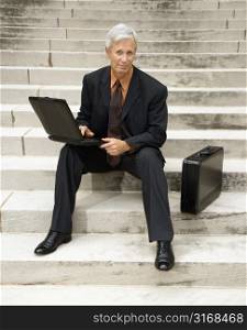 Caucasian middle aged businessman sitting on steps outdoors with laptop and briefcase looking at viewer.
