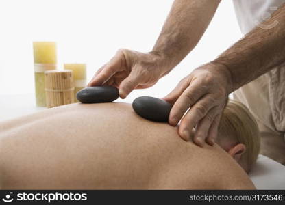 Caucasian middle-aged male massage therapist placing hot stones on back of Caucasian middle-aged woman lying on massage table.