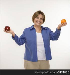 Caucasian middle aged woman holding fruit in outstretched arms and smiling at viewer.