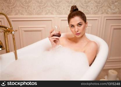 Caucasian nude woman with glass of red wine in the bathtub with foam.