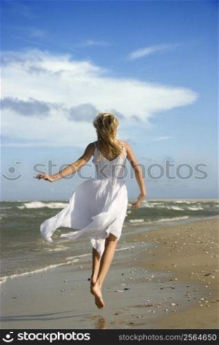 Caucasian pre-teen girl running down the beach in flowing white dress.