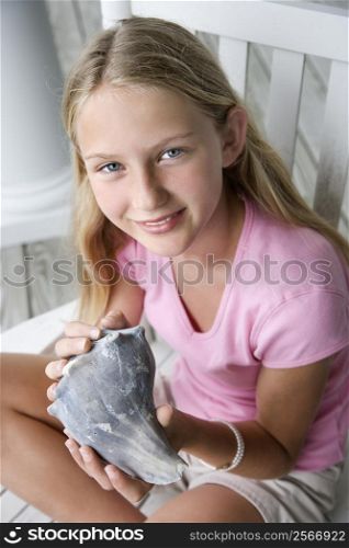 Caucasian pre-teen girl sitting indian style in rocking chair holding conch shell looking at viewer and smiling.
