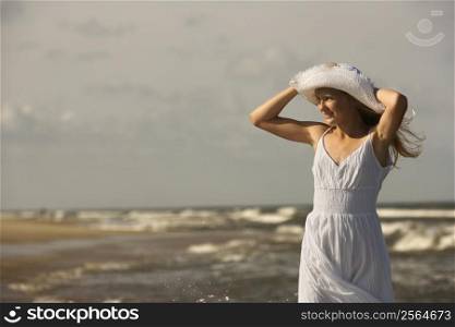 Caucasian pre-teen girl standing on beach holding hat on head.