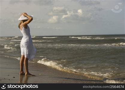 Caucasian pre-teen girl standing on beach holding hat on head.