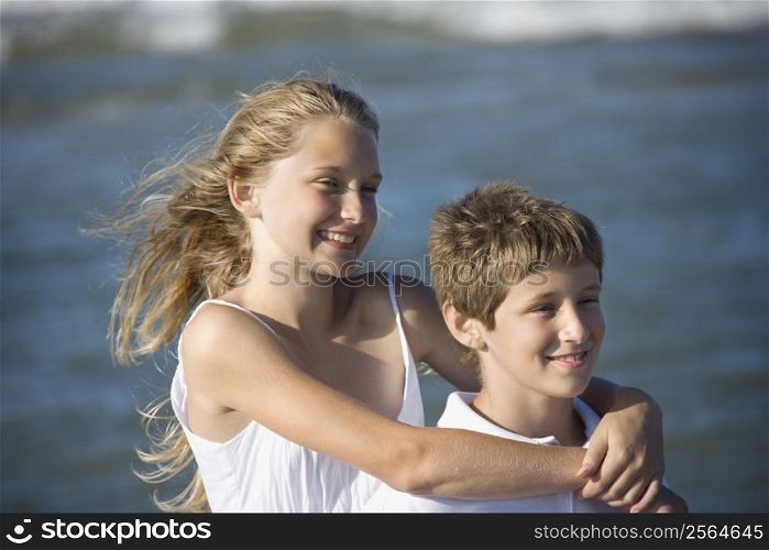 Caucasian pre-teen girl with arms around pre-teen boy on beach.