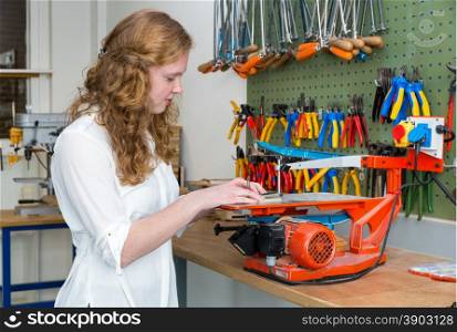 Caucasian redhead teenage girl operating electric jigsaw in classroom of school building