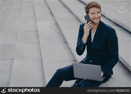 Caucasian satisfied man in formal outfit sitting on steps outdoor, talking on mobile phone and working on laptop computer. Male freelancer typing on keyboard while speaking on cellphone with client. Caucasian bearded young man sitting on steps outdoor with laptop and talking on mobile phone