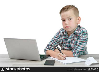 Caucasian school-age boy in a plaid shirt sitting at the table and writing in a piece of paper . there is also a laptop and a phone on the table