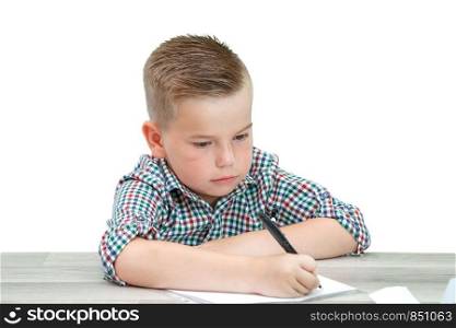 Caucasian school-age boy in a plaid shirt sitting at the table and writing in a piece of paper . there is also a laptop and a phone on the table