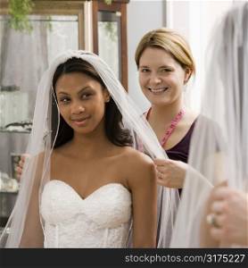 Caucasian seamstress helping African-American bride with veil in bridal shop.