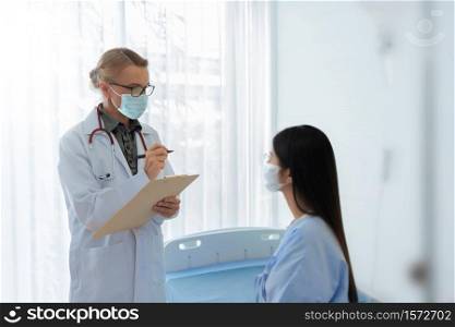 Caucasian Senior female doctor explaining and giving advice to Asian patient in medical room together. Mature physician and Young woman wear protective face masks talking together in hospital.