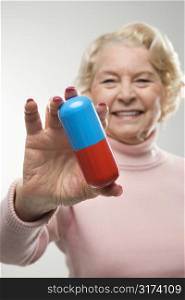Caucasian senior woman holding oversized pill at viewer.