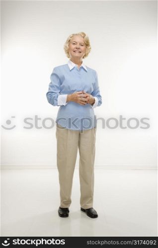 Caucasian senior woman standing with hands together smiling at viewer.