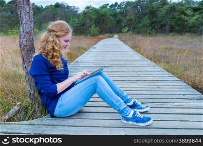 Caucasian teenage girl working with tablet computer on wooden footpath in nature