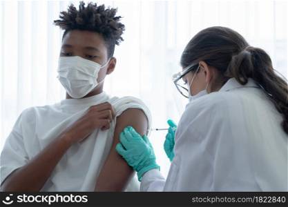 Caucasian woman doctor wear mask, face shield and gloves is giving injections or vaccines to arm of African American boy at hospital. Preventing spread of COVID-19 by vaccinating people concept
