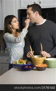 Caucasian woman feeding man at kitchen counter while he makes salad.