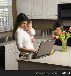 Caucasian woman holding baby and typing on laptop computer in kitchen.
