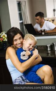 Caucasian woman hugging toddler son in kitchen with father on laptop in background.