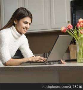 Caucasian woman in kitchen looking at laptop computer.