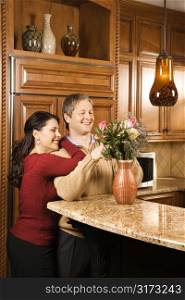 Caucasian woman leaning on Caucasian man while he arranges flowers in vase in kitchen.
