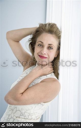 Caucasian woman leaning on wall smiling at viewer with hands touching head.