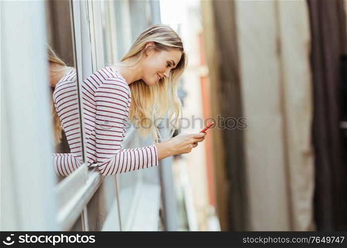 Caucasian woman leaning out of her house window using a smart phone.. Caucasian woman leaning out of her house window using a smartphone.