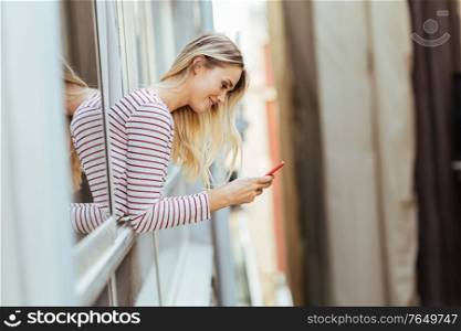 Caucasian woman leaning out of her house window using a smart phone.. Caucasian woman leaning out of her house window using a smartphone.