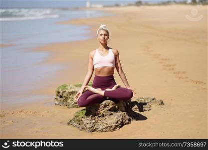 Caucasian woman practicing yoga at seashore. Young female doing lotus pose in the beach in Cadiz, Andalusia, Spain.. Caucasian blonde woman practicing yoga in the beach