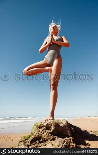 Caucasian woman practicing yoga at seashore. Young female standing on one leg while practicing the tree pose on a tranquil beach in Cadiz, Andalusia, Spain.. Caucasian blonde woman practicing yoga in the beach