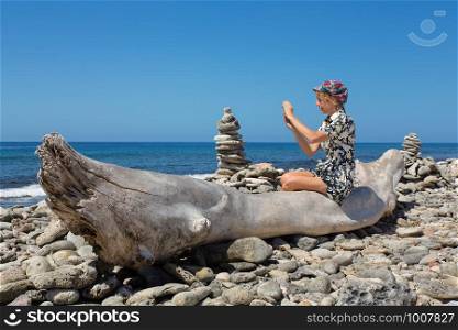 Caucasian woman sitting on tree trunk photographing at coast