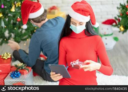 Caucasian woman with hygiene mask choose the gift from online shop and ask help from her couple for celebration of Christmas during Covid-19 pandemic and new normal lifestyle to stay at home.